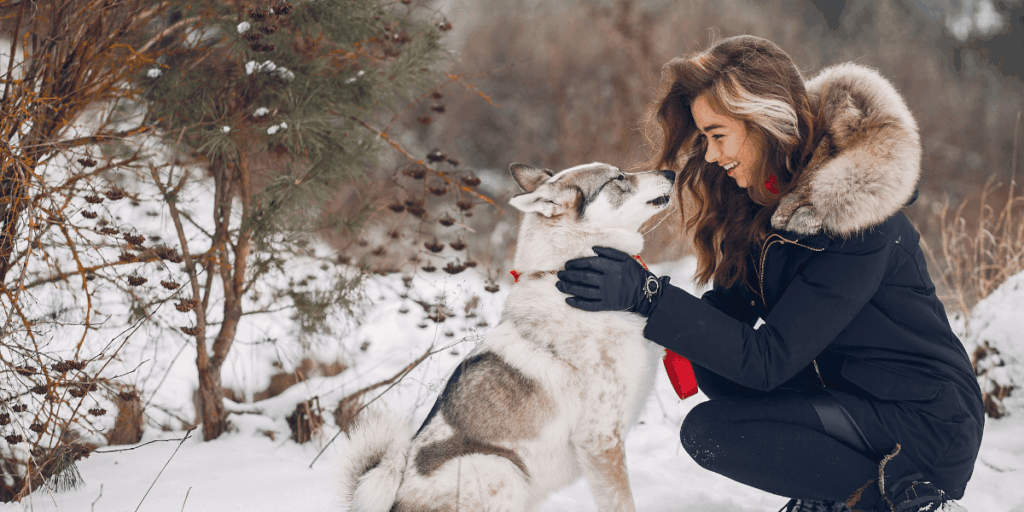 A woman kneels in the snow, playfully interacting with her dog beside her.
