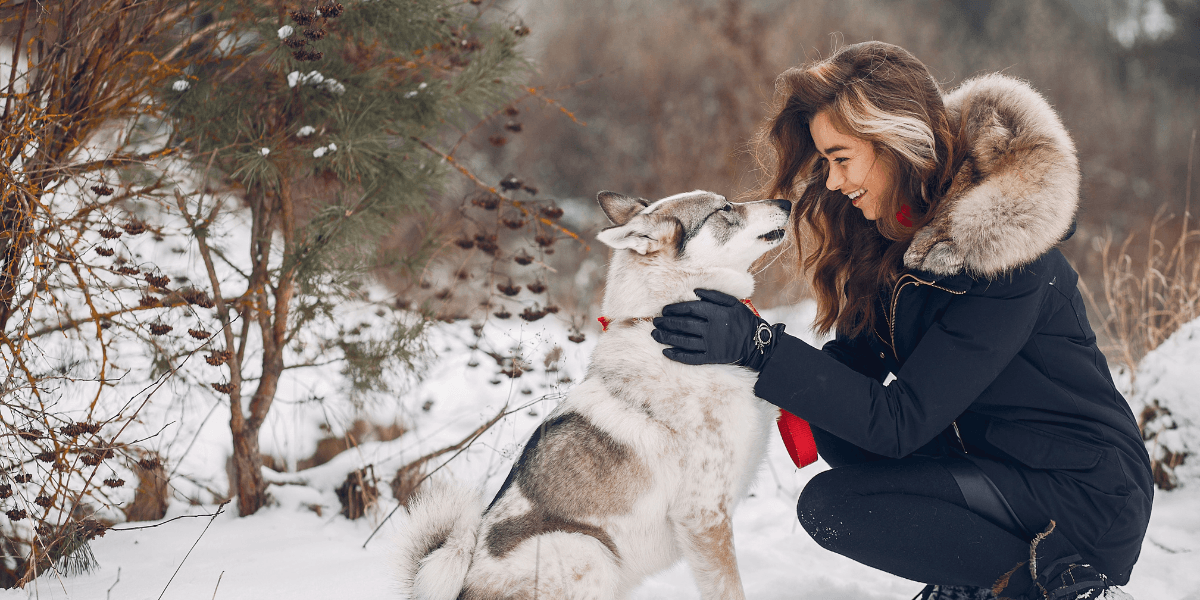 A woman kneels in the snow, playfully interacting with her dog beside her.
