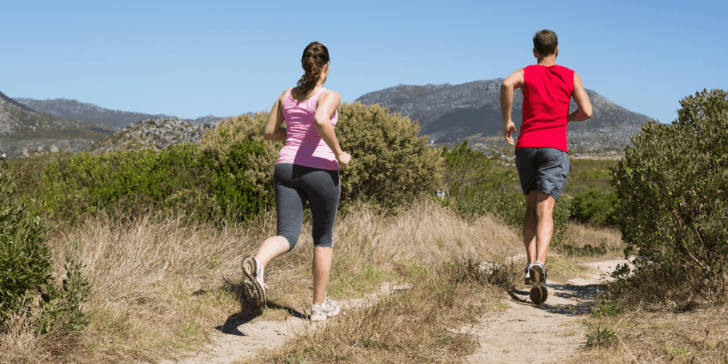  A man and woman running side by side on a dirt trail surrounded by trees and greenery.