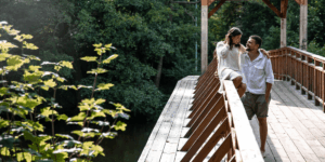 A couple enjoys a peaceful moment on a wooden bridge, framed by dense woods and vibrant greenery.