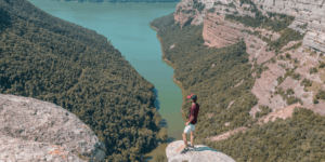 A man stands on a cliff's edge, gazing out over a serene lake surrounded by lush greenery.