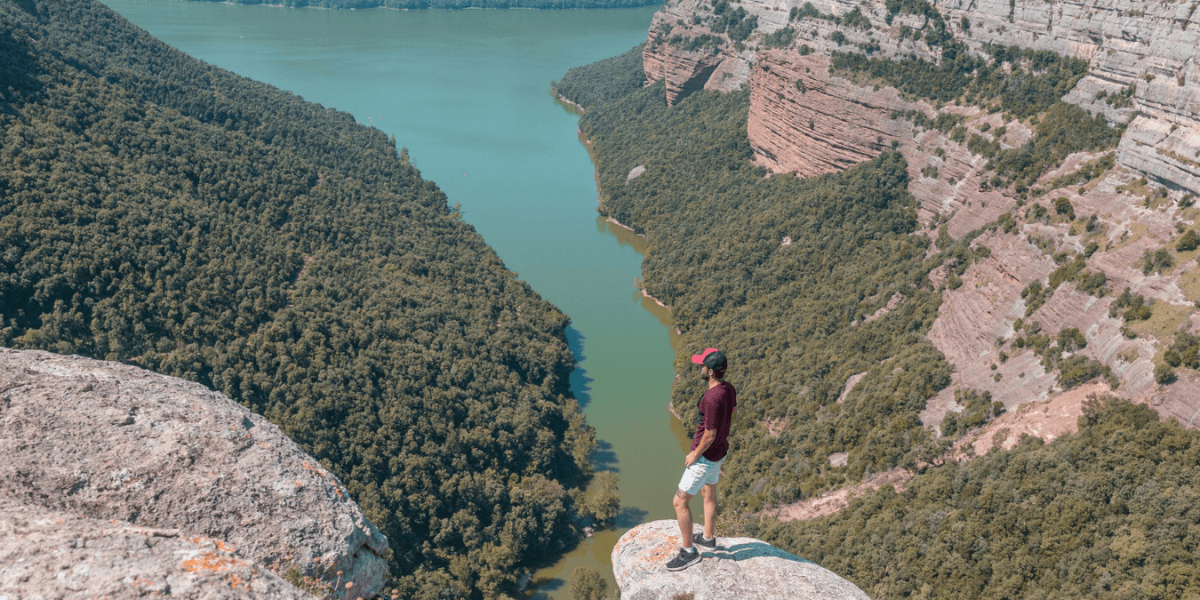 A man stands on a cliff's edge, gazing out over a serene lake surrounded by lush greenery.
